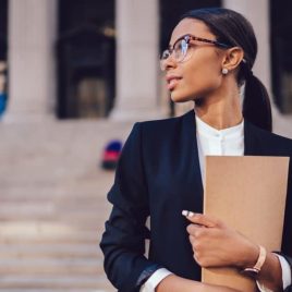 Pensive African American female lawyer in stylish formal suit holding folder with mock up area and looking away standing against courthouse. Half length of woman professional advocate with documents; Shutterstock ID 1008711700; Notes: Muse.com